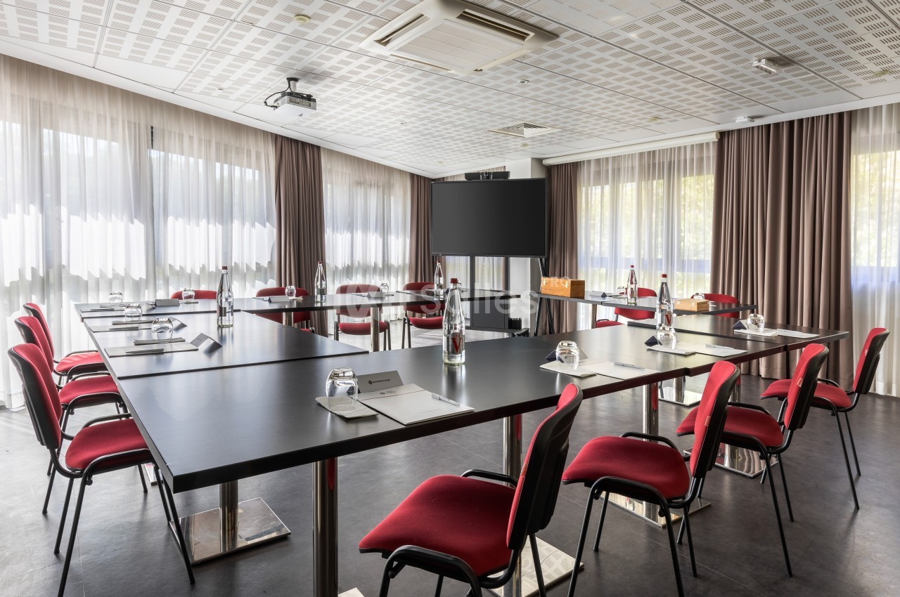 Salle de réunion lumineuse avec tables noires disposées en U, chaises rouges et écran de présentation.
