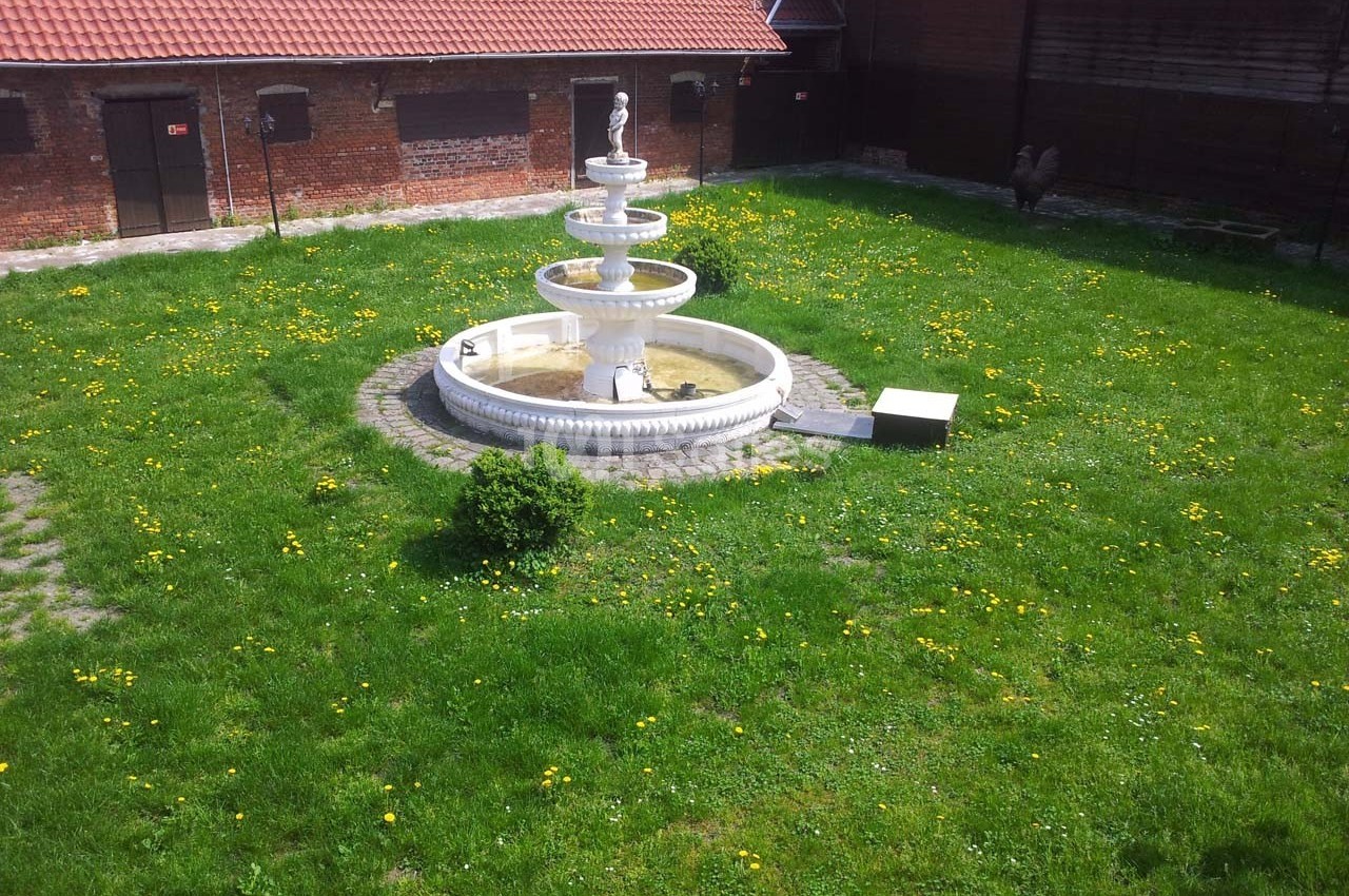 Fontaine blanche à trois niveaux entourée d'une pelouse verte parsemée de pissenlits, dans une cour bordée de bâtiments en…