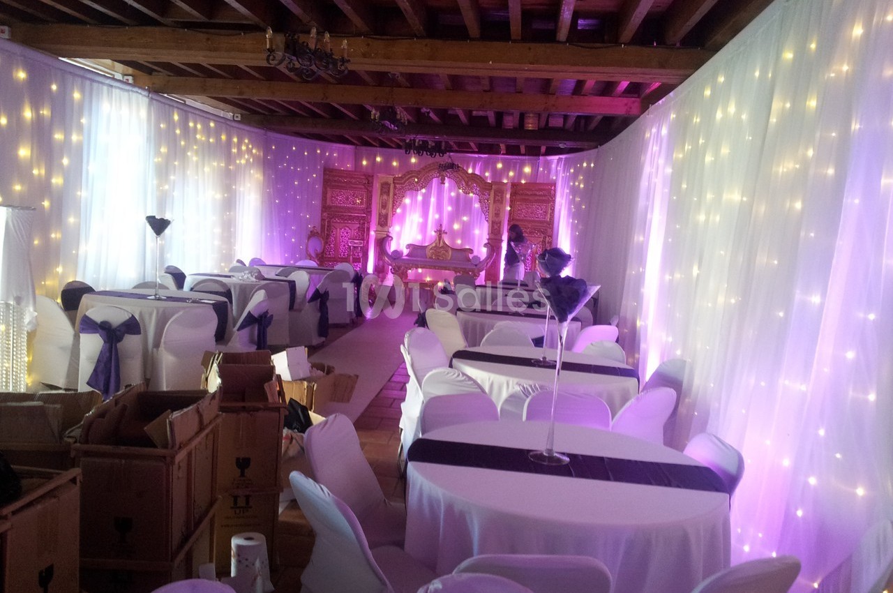 Salle décorée pour un événement avec des tables drapées de blanc, des lumières féeriques et un espace central aménagé.