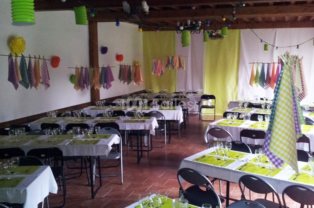 Salle décorée pour un événement, avec des tables dressées, nappes blanches et serviettes colorées suspendues.