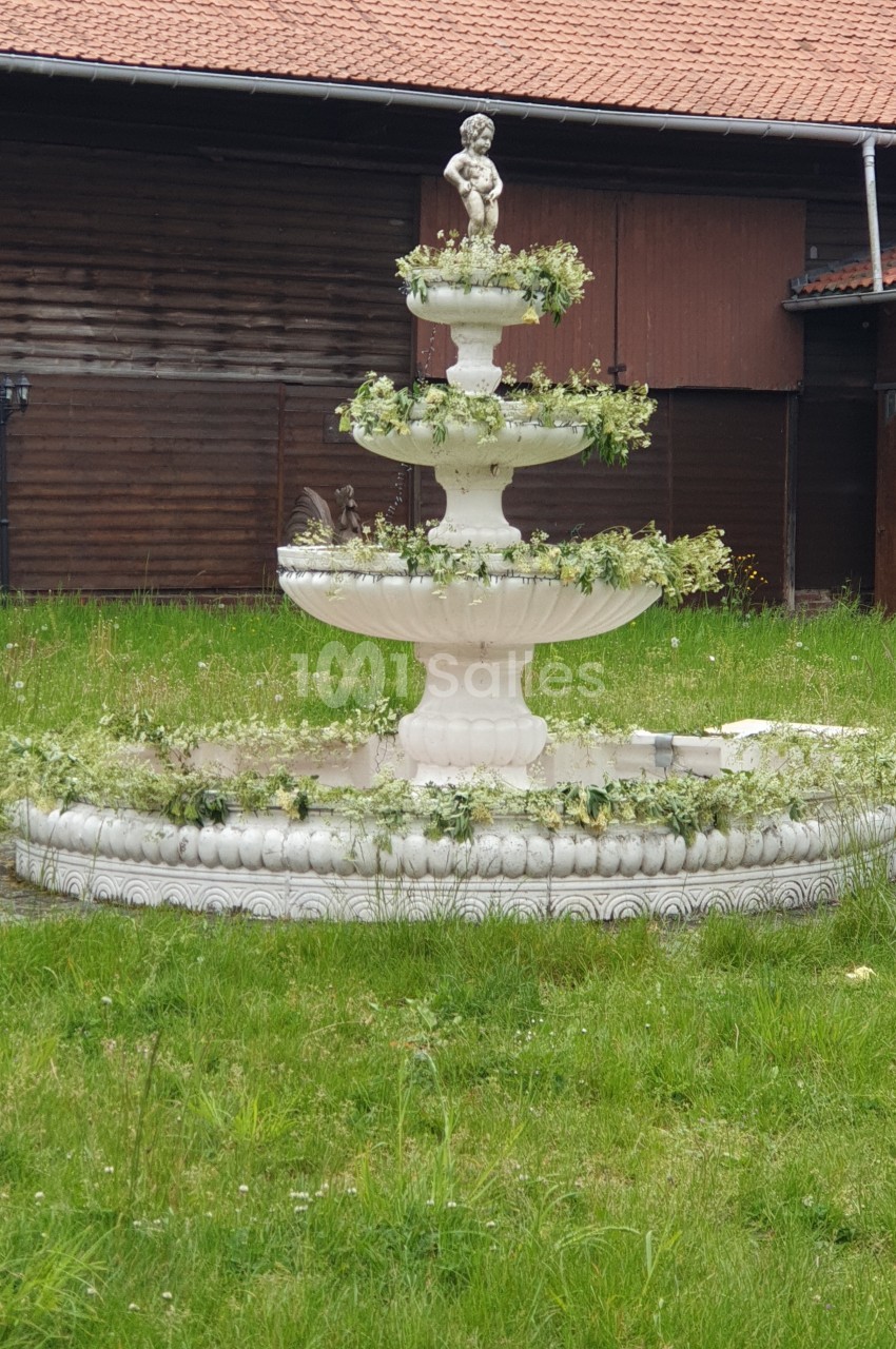 Fontaine blanche à trois niveaux ornée de végétation, avec une statue d'enfant au sommet, située sur une pelouse devant un…