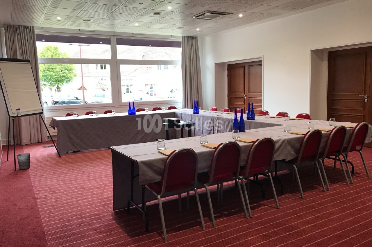 Salle de réunion lumineuse avec tables disposées en U, chaises rouges, tableau de conférence et bouteilles d'eau bleues.