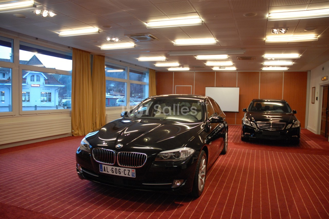 Deux voitures noires exposées dans une salle éclairée avec moquette rouge et grandes fenêtres donnant sur l'extérieur.