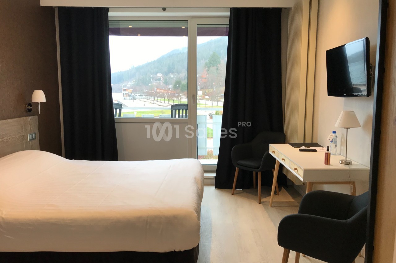 Chambre d'hôtel lumineuse avec lit double, bureau, deux fauteuils, télévision murale et grande fenêtre donnant sur un…
