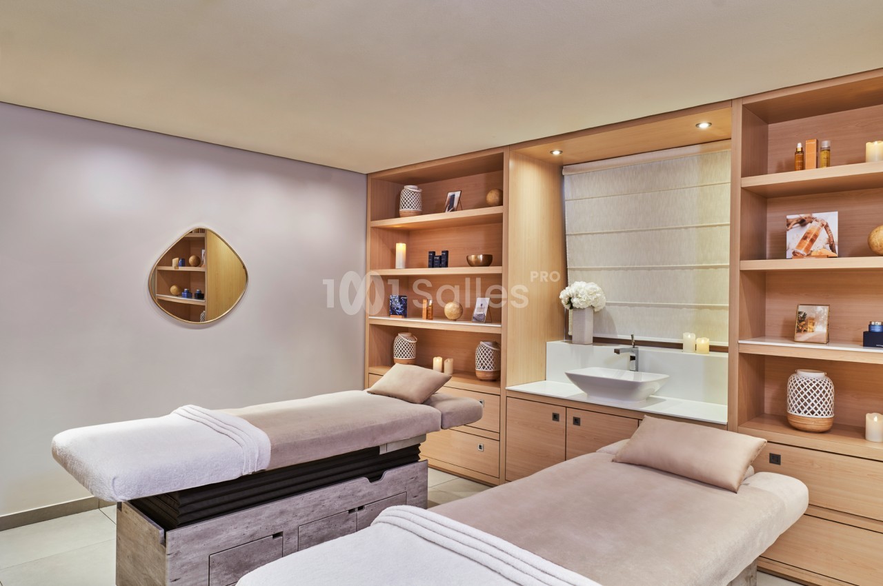 Deux tables de massage dans une salle lumineuse avec étagères en bois, décorée de bougies et d'objets apaisants.