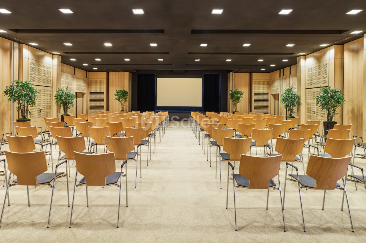 Salle de conférence moderne avec rangées de chaises en bois, écran de projection et plantes décoratives.