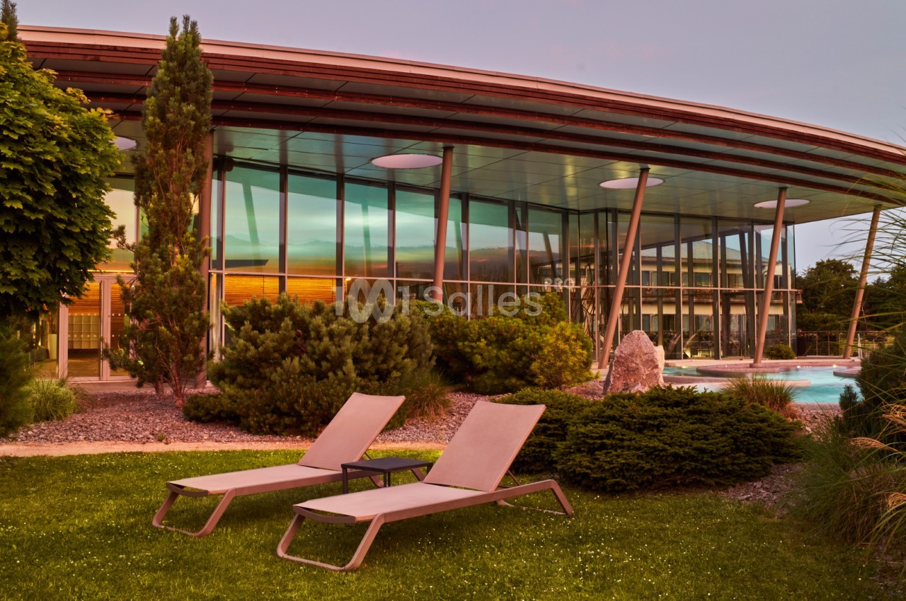 Façade vitrée d'un bâtiment moderne entouré de végétation, avec deux chaises longues sur une pelouse au premier plan.