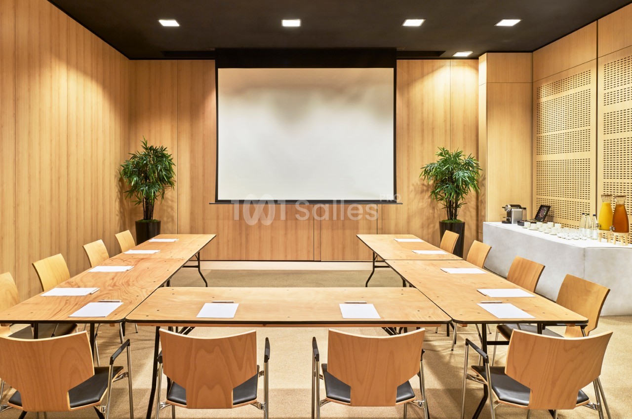 Salle de réunion avec tables en U, chaises en bois, écran de projection, plantes et buffet avec boissons.