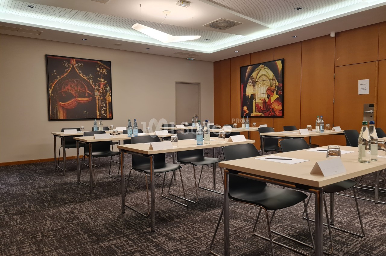 Salle de réunion avec tables disposées en rangées, chaises noires, bouteilles d'eau et œuvres d'art accrochées aux murs.