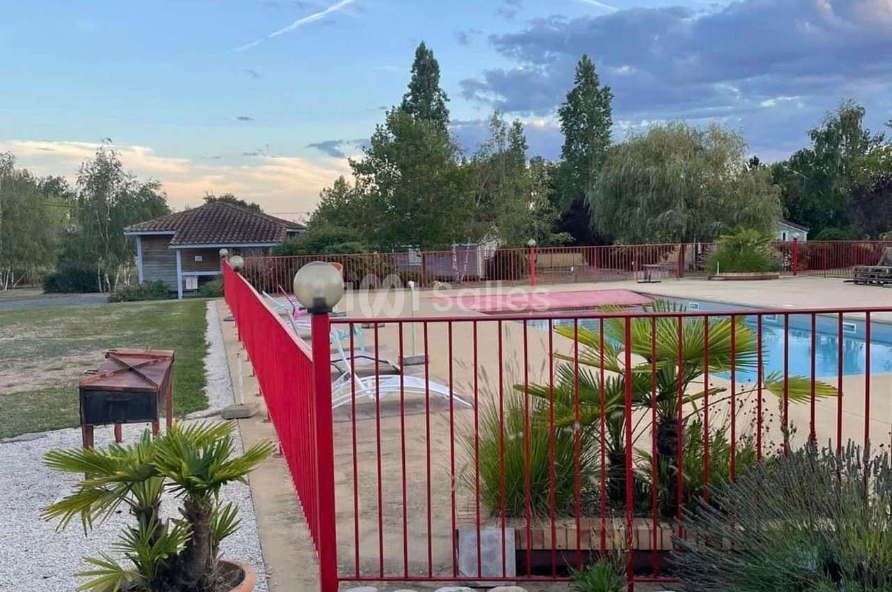 Clôture rouge entourant une piscine extérieure avec transats, arbres et ciel nuageux en arrière-plan.