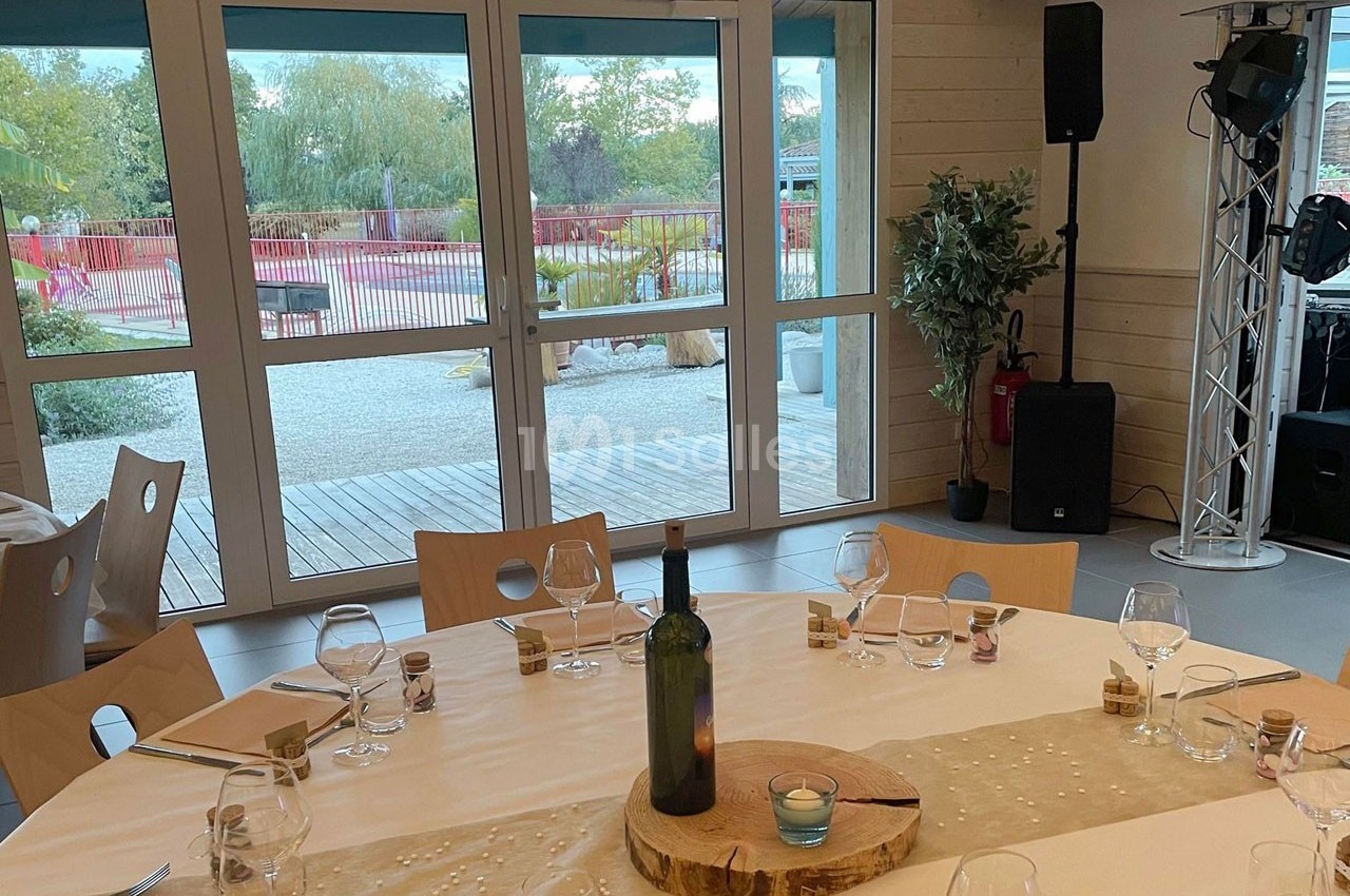 Table dressée avec nappage beige, verres et décoration en bois, devant une baie vitrée donnant sur un espace extérieur.