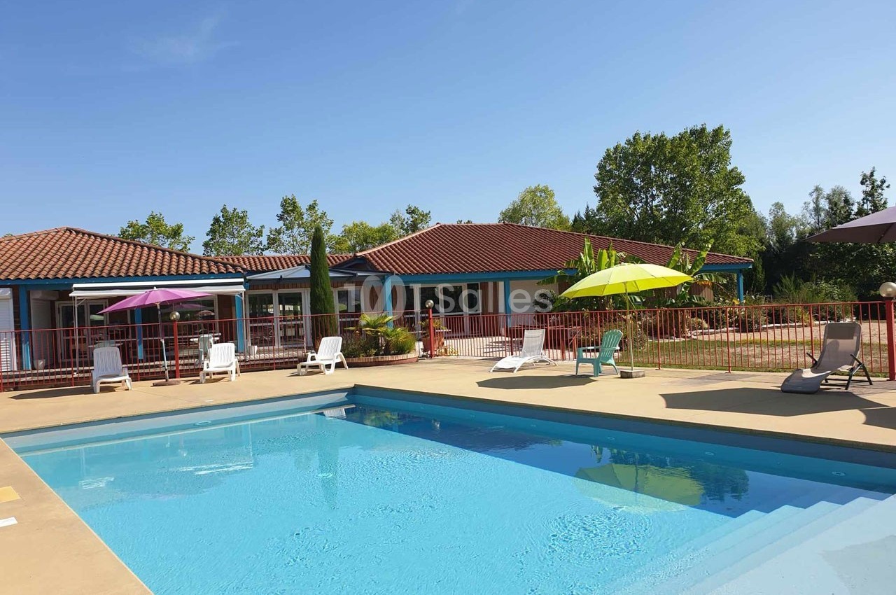 Piscine extérieure entourée de transats et parasols colorés, avec un bâtiment à toit rouge en arrière-plan.