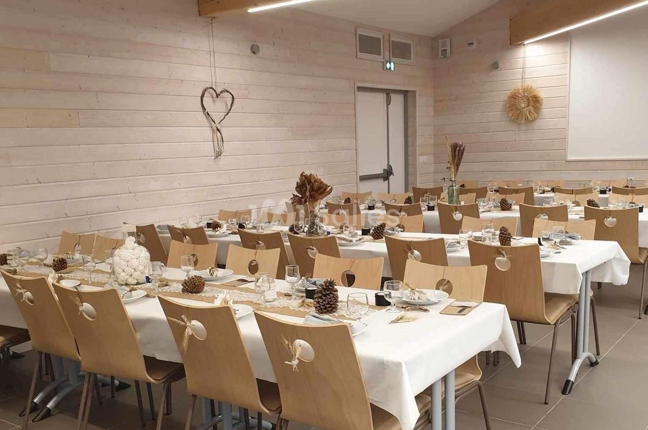 Salle décorée pour un repas, avec des tables alignées, nappes blanches, décorations naturelles et chaises en bois.