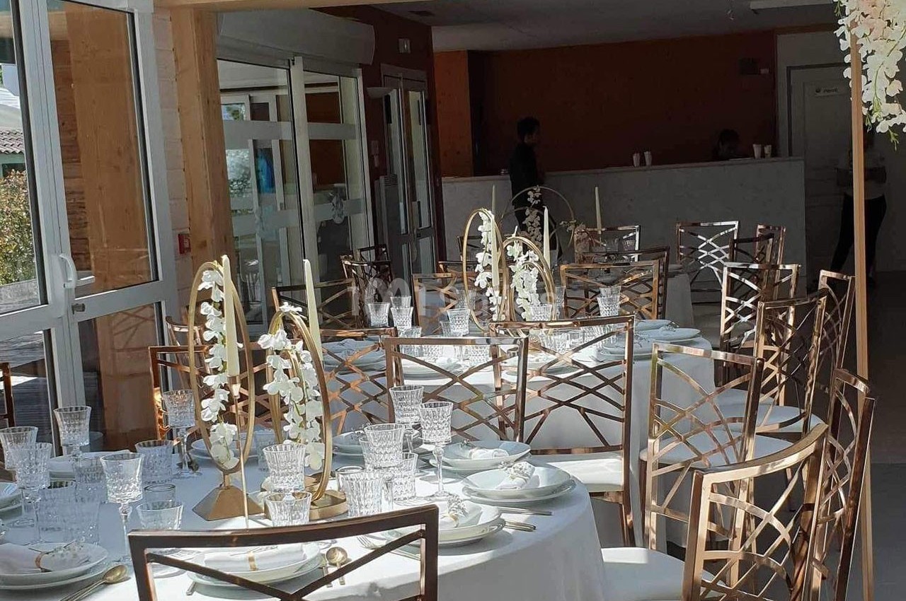 Salle lumineuse avec tables rondes dressées, décorées de fleurs blanches et bougies, prête pour un événement.