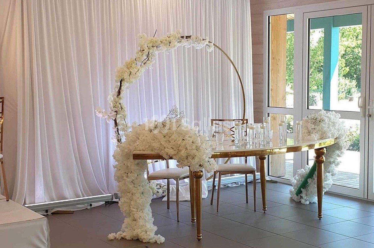 Table décorée pour un événement, ornée de fleurs blanches et d'une arche circulaire, devant un rideau blanc.