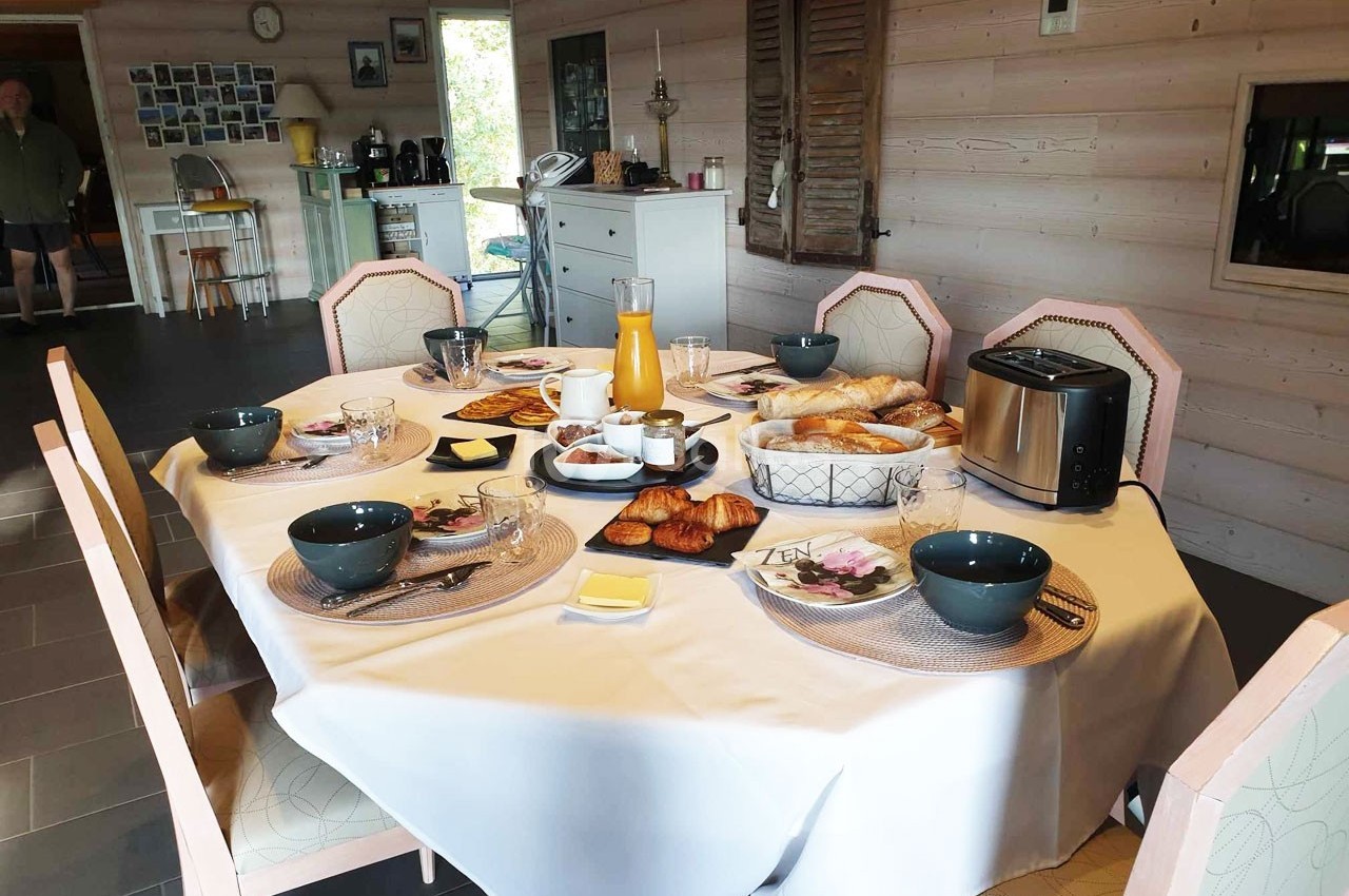 Table dressée pour un petit-déjeuner avec viennoiseries, pain, jus d'orange et vaisselle dans une salle à manger lumineuse.