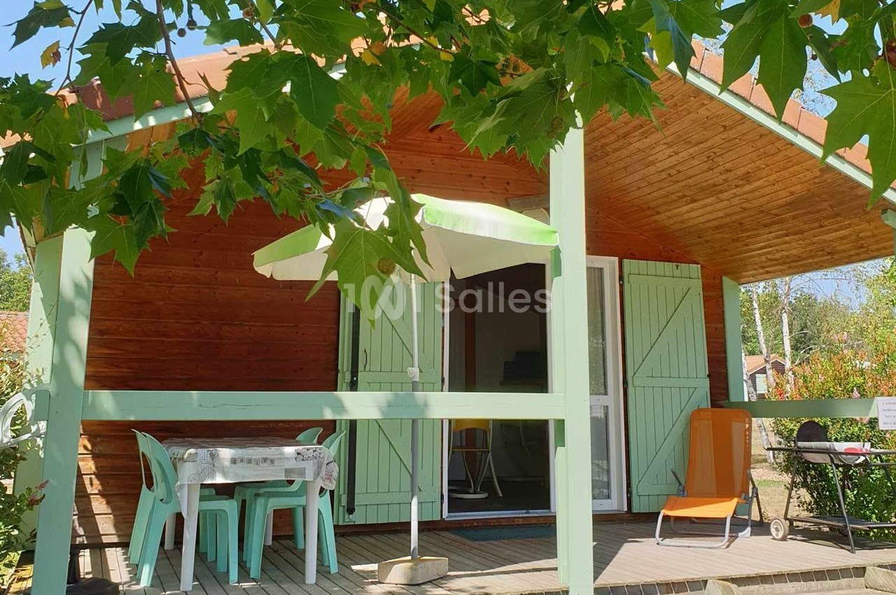 Chalet en bois avec terrasse ombragée, table, chaises, parasol et transat, entouré de verdure.
