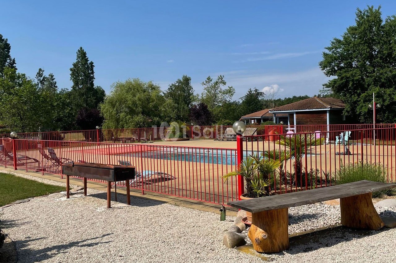 Piscine extérieure entourée d'une barrière rouge, avec un banc en bois et un barbecue au premier plan.