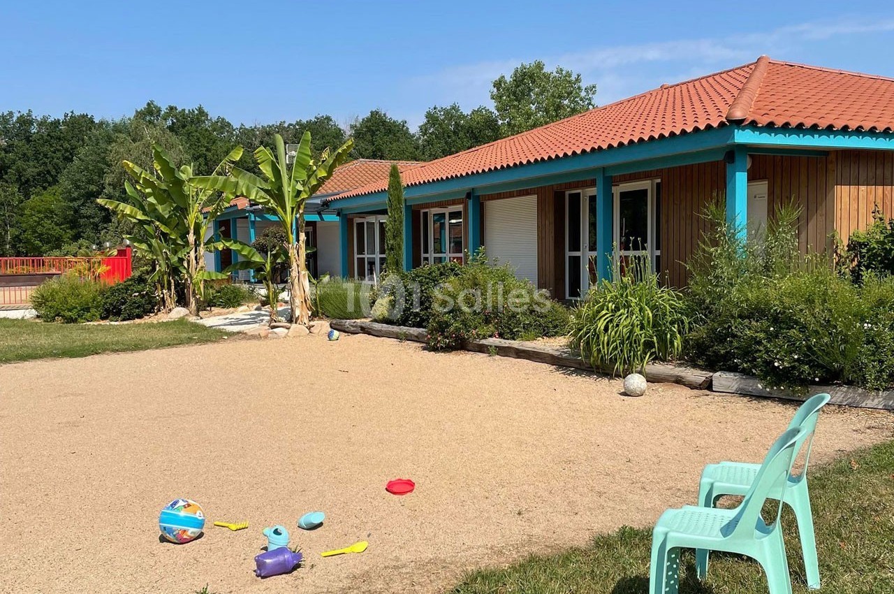 Cour extérieure d'une maison avec terrasse, plantes tropicales, jouets pour enfants au sol et chaises en plastique.