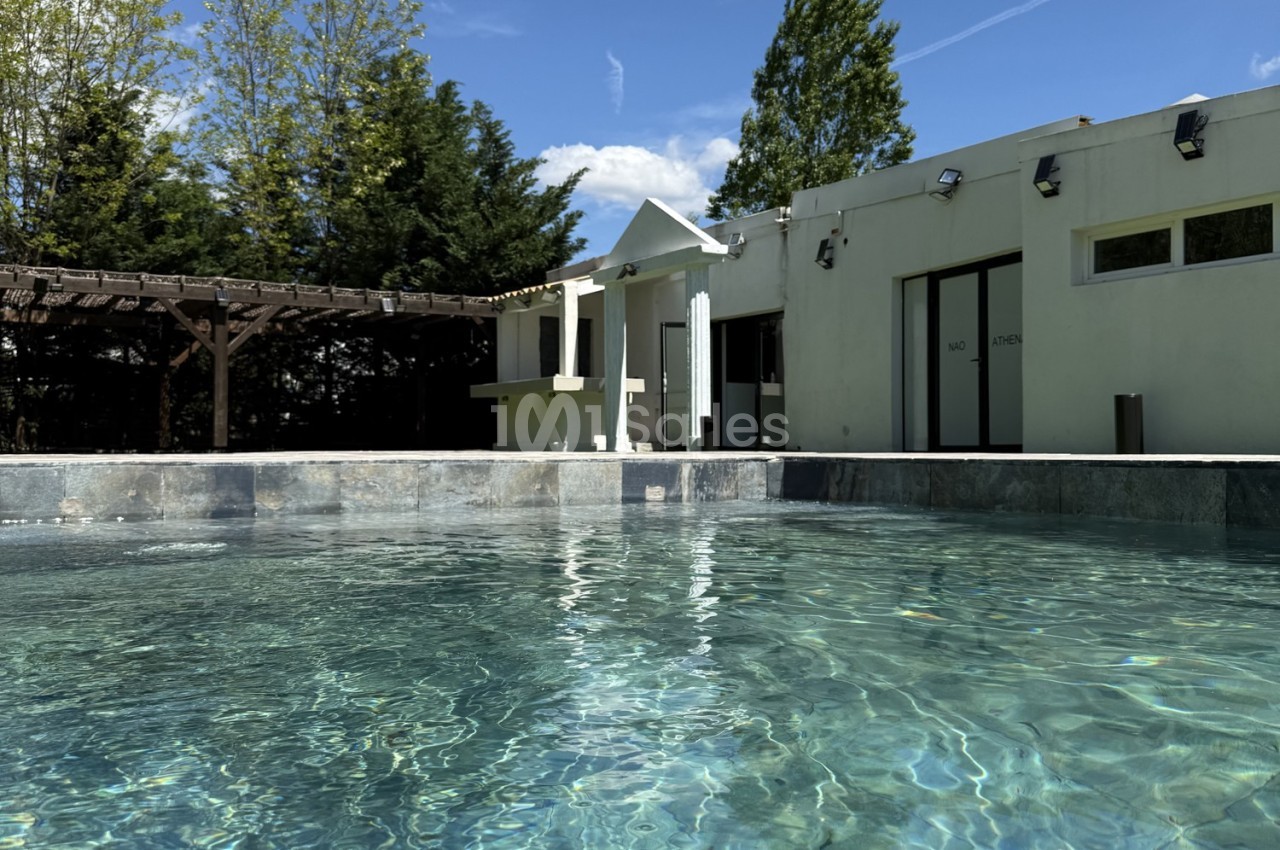 Piscine extérieure avec eau claire, entourée d'une terrasse, d'arbres et d'un bâtiment moderne sous un ciel dégagé.