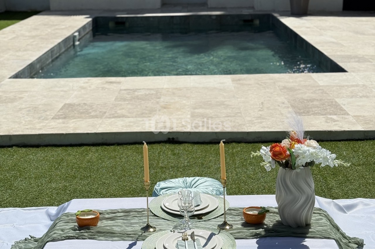Table dressée avec vaisselle élégante, bougies et fleurs, disposée devant une piscine entourée de pierre et d'herbe.
