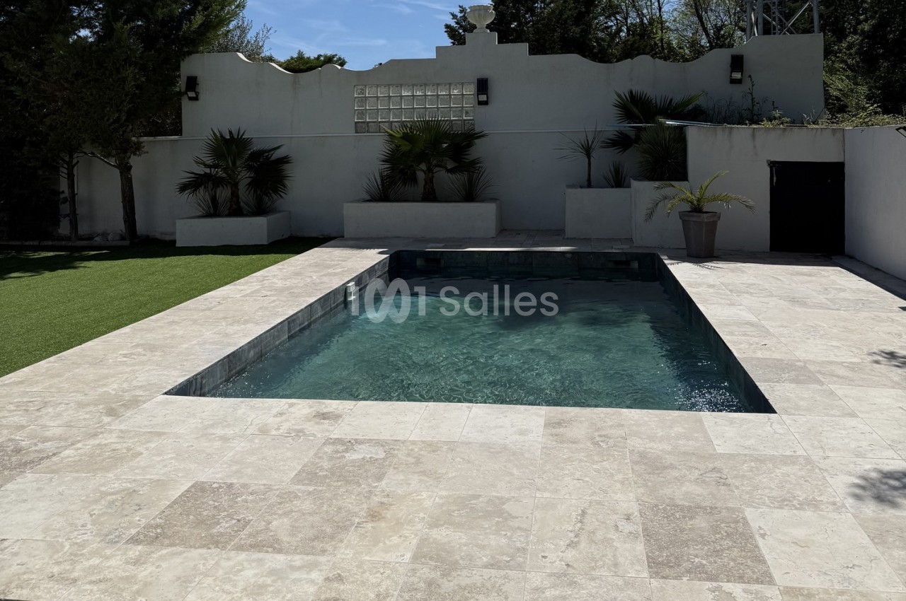 Piscine rectangulaire entourée de dalles claires, avec des plantes et un mur blanc en arrière-plan.