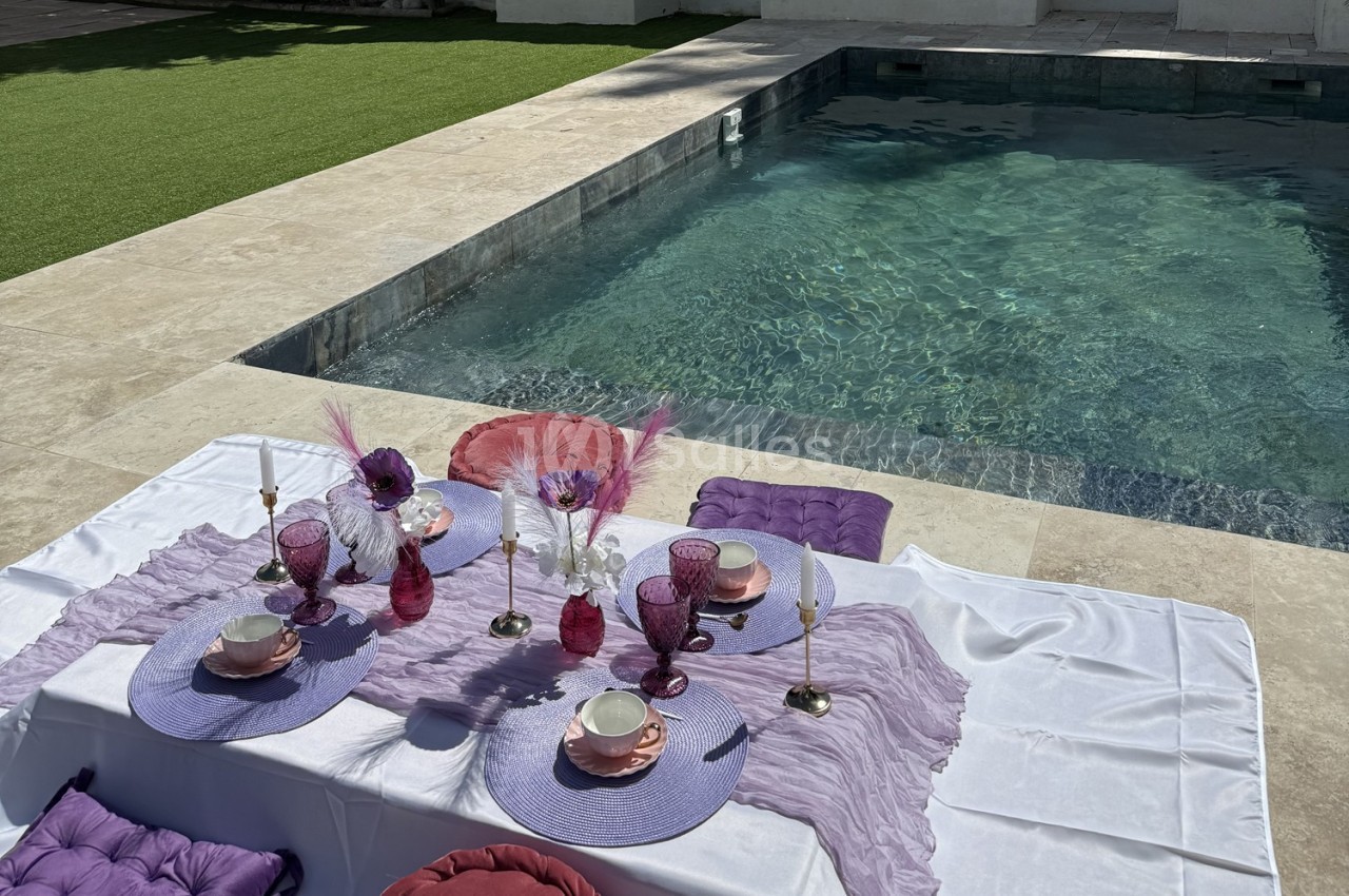 Table dressée avec vaisselle rose et violette, décorée de plumes et fleurs, près d'une piscine et d'une pelouse.