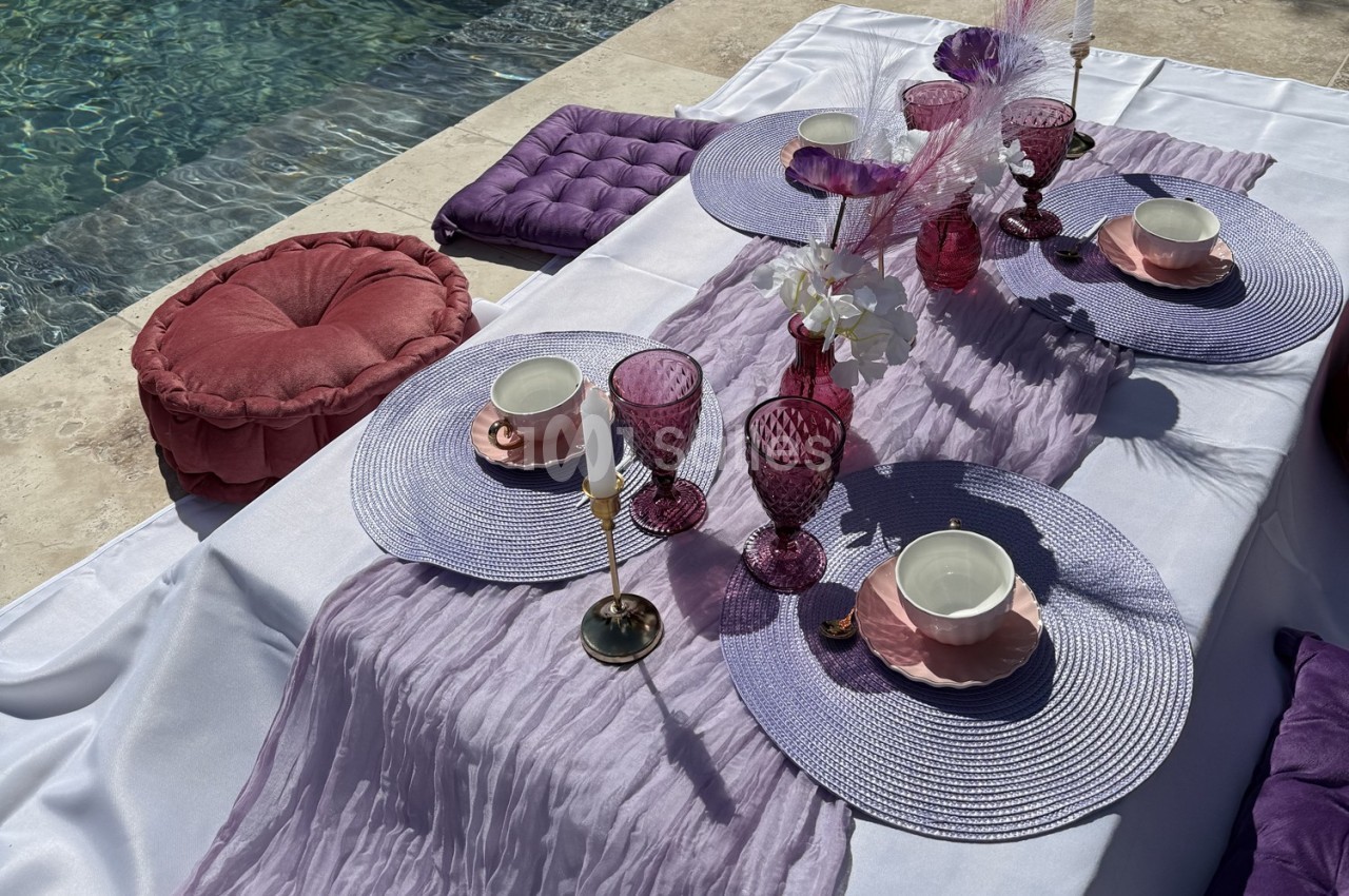 Table dressée en extérieur avec nappes blanches, décorations violettes, verres roses et coussins près d'une piscine.