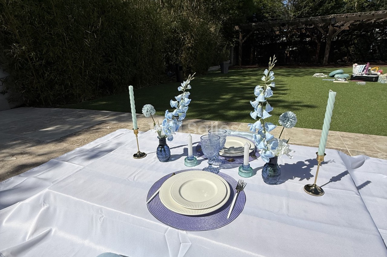 Table extérieure dressée avec vaisselle blanche, bougies et fleurs bleues, sur une nappe blanche dans un jardin.
