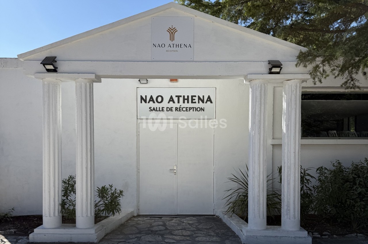 Façade blanche avec colonnes et porte centrale indiquant ’Nao Athena - Salle de réception’, entourée de végétation.