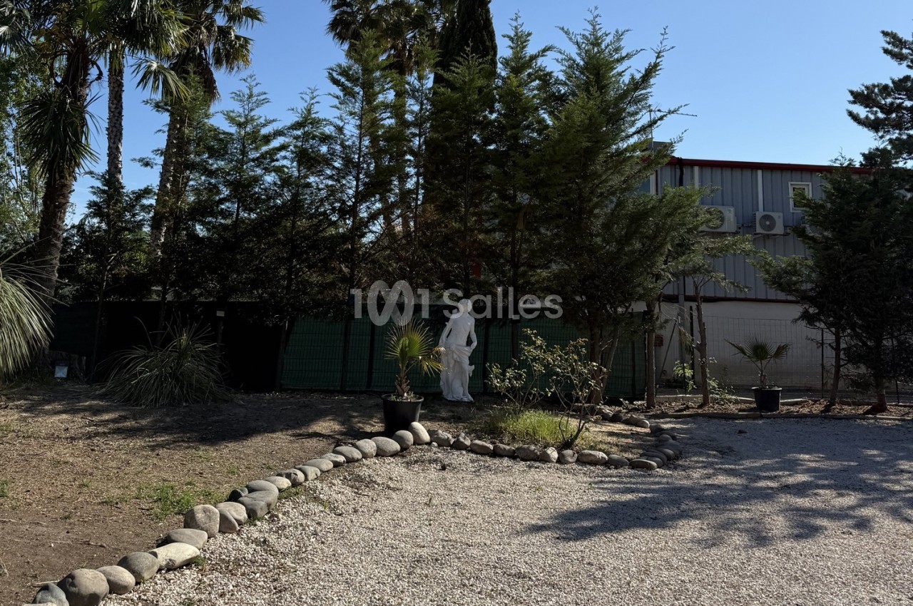 Allée gravillonnée bordée de pierres, avec une statue blanche entourée d'arbres et un bâtiment en arrière-plan.