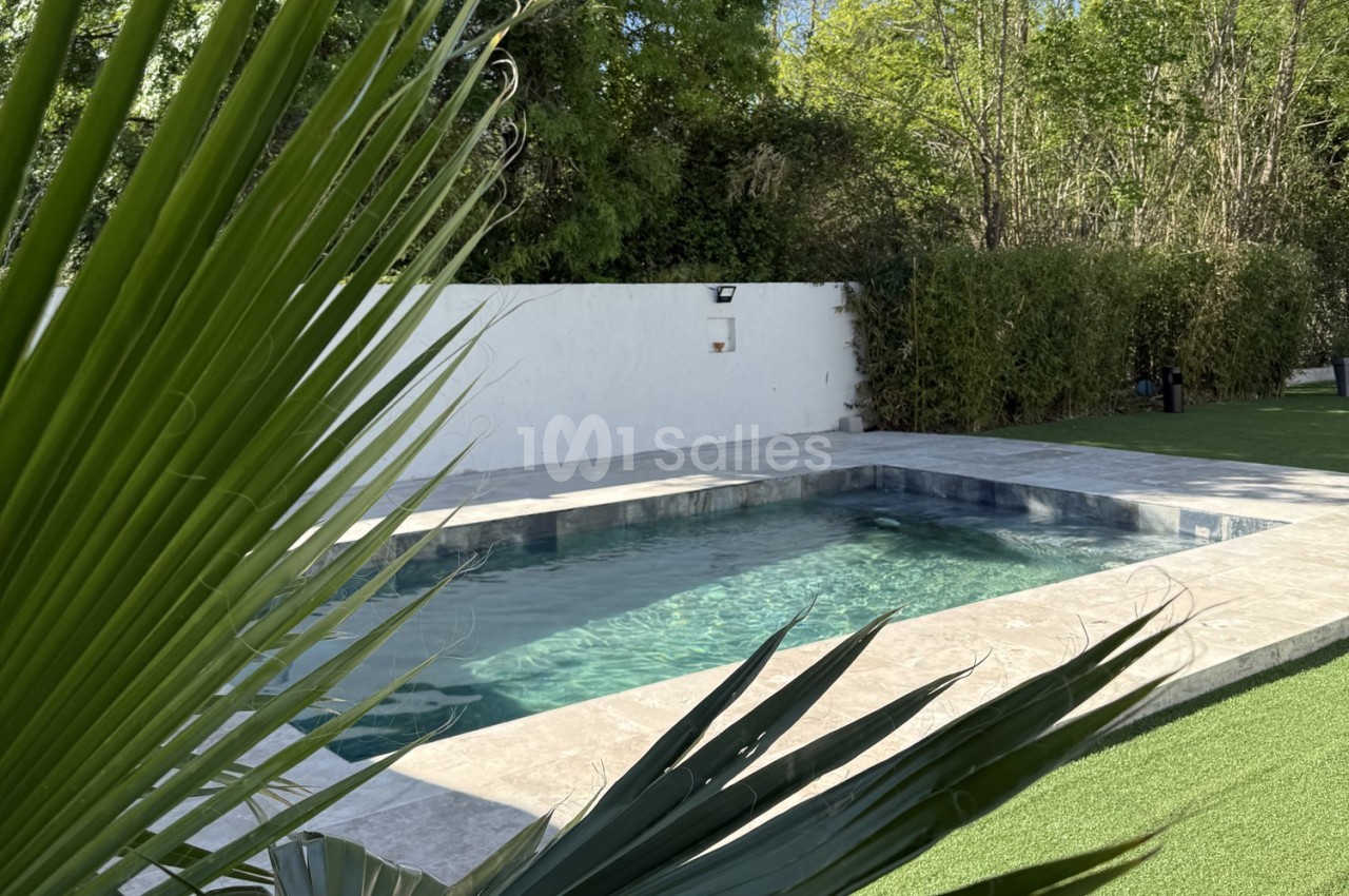 Piscine extérieure entourée de dalles en pierre, avec pelouse artificielle, haies et arbres en arrière-plan.