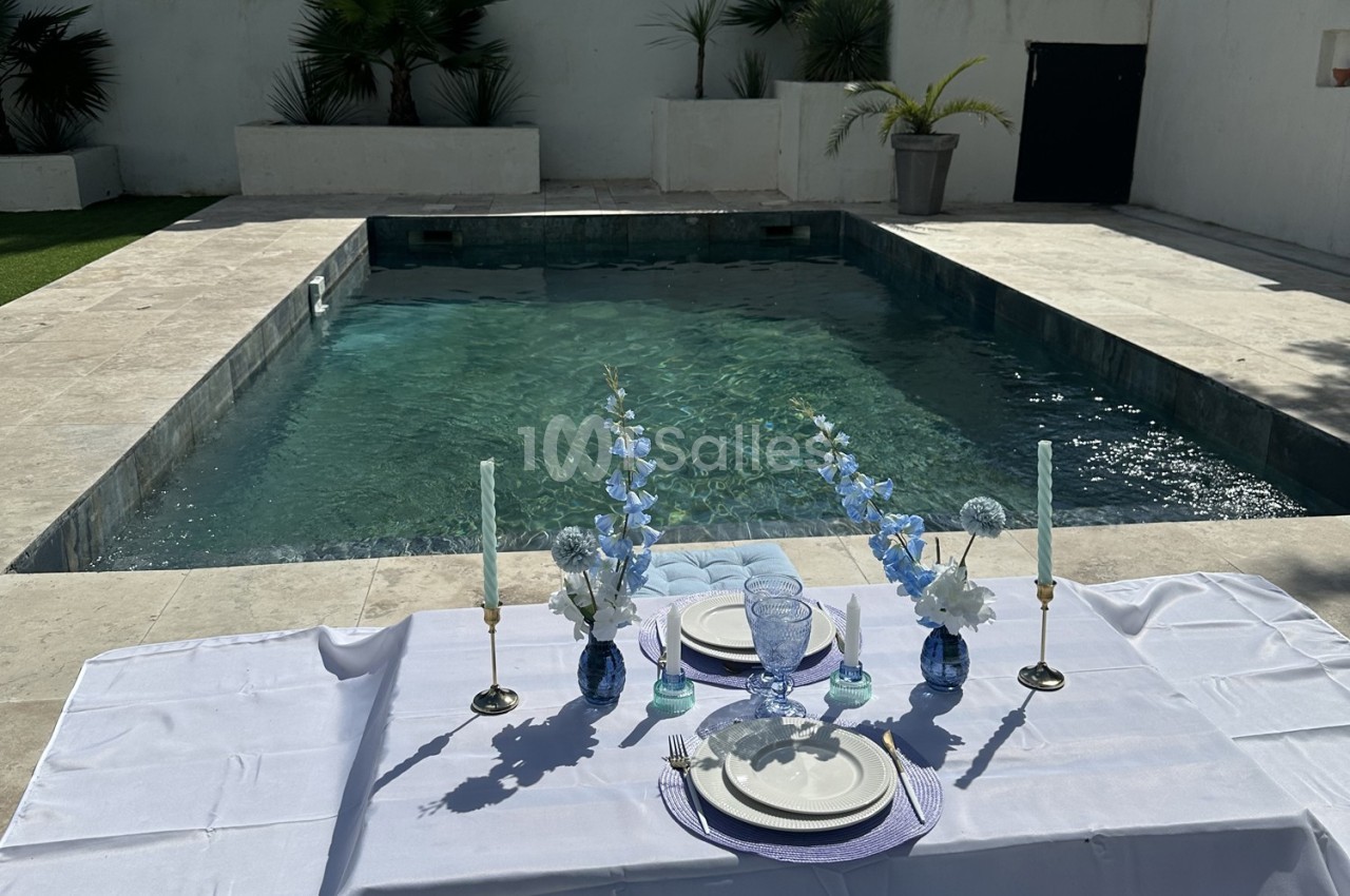 Table dressée avec vaisselle et fleurs bleues près d'une piscine entourée de dalles claires et de végétation.
