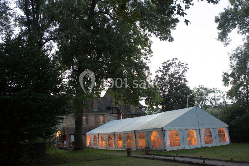 Grande tente blanche éclairée de l'intérieur, installée dans un jardin arboré près d'une maison en pierre au crépuscule.