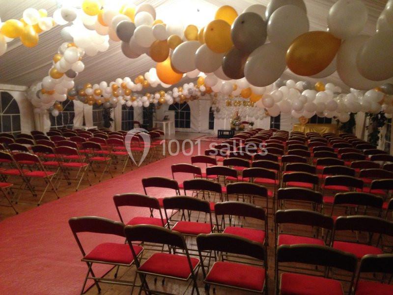 Salle décorée avec des ballons blancs et dorés, rangées de chaises rouges alignées sur un tapis rouge.