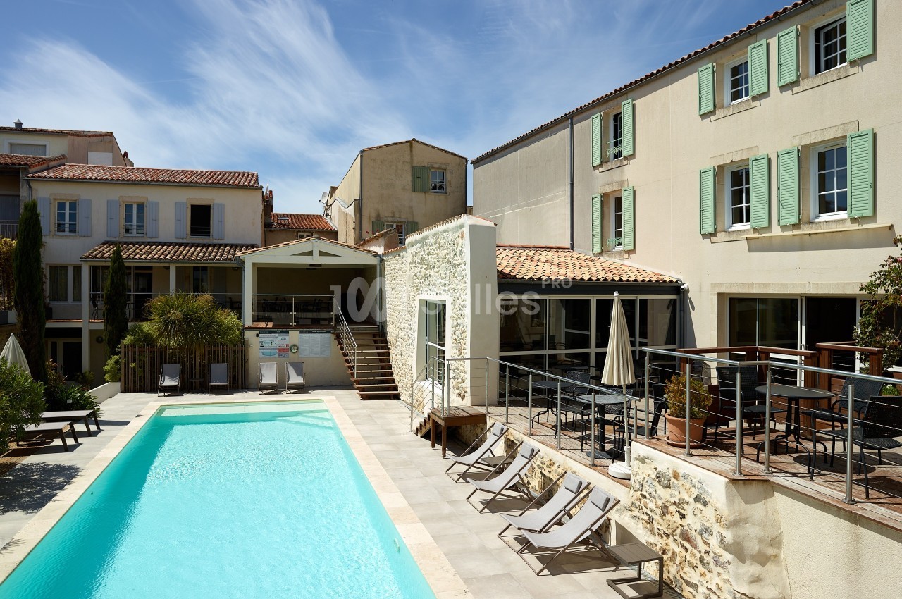 Piscine extérieure entourée de chaises longues et bâtiments aux façades claires sous un ciel dégagé.