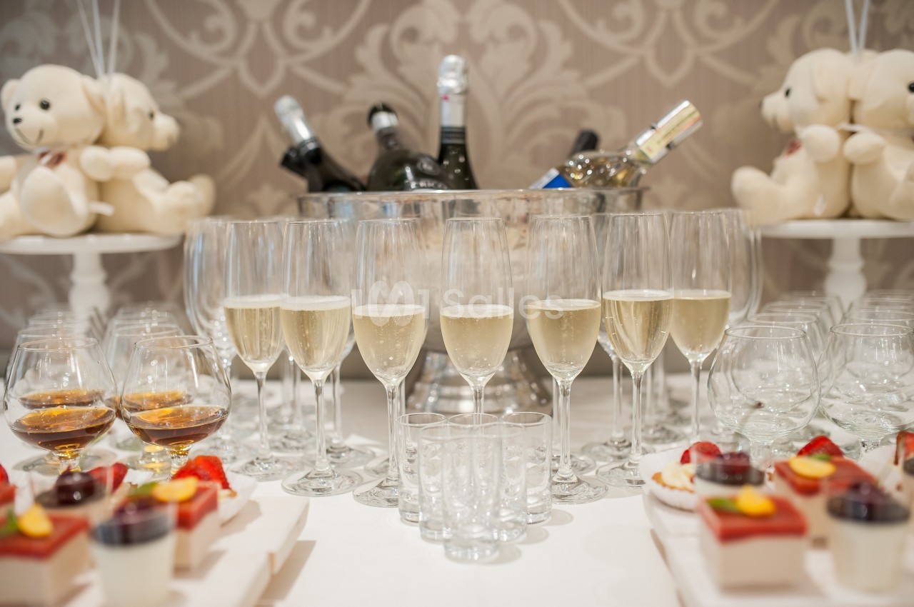 Verres de champagne alignés devant un seau à glace rempli de bouteilles, avec desserts et peluches en arrière-plan.