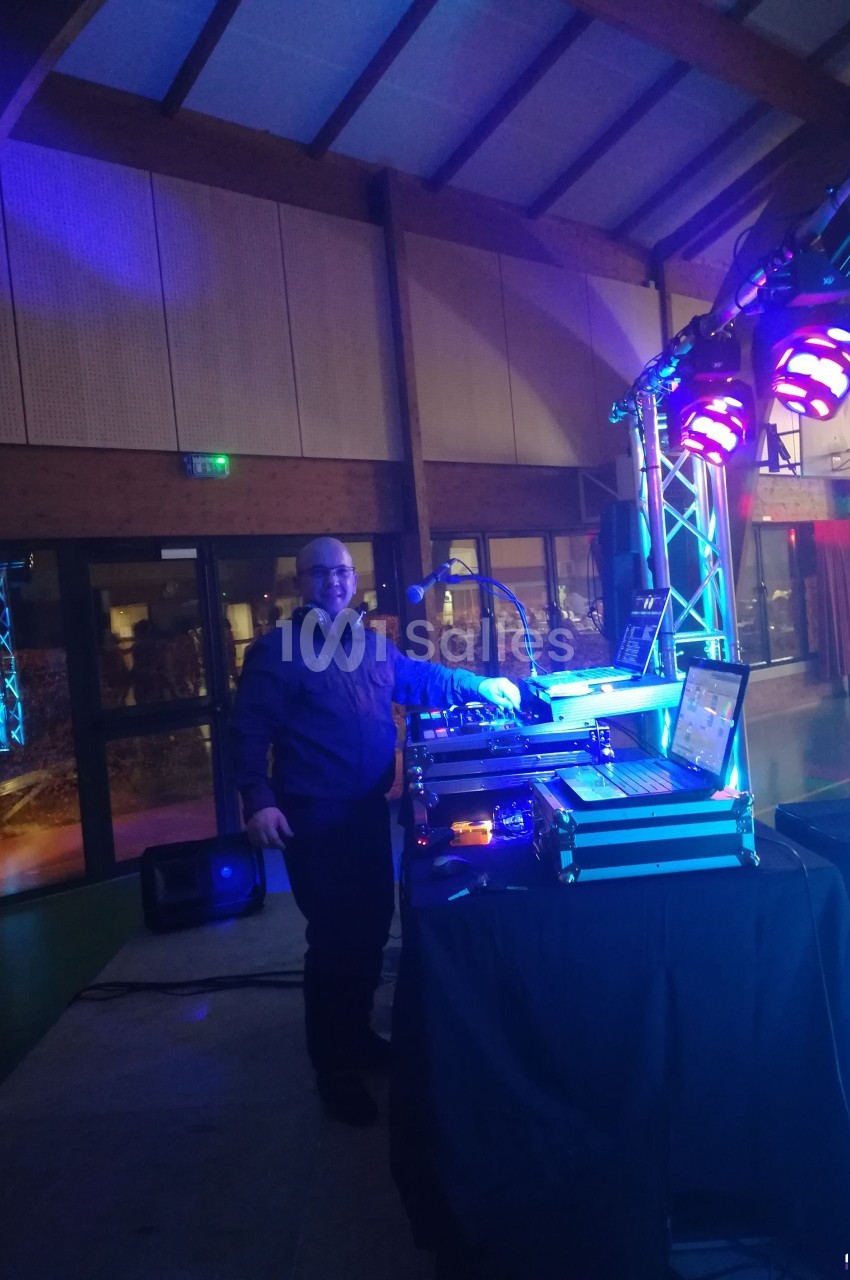 Un DJ souriant se tient derrière une table de mixage éclairée par des lumières colorées dans une salle de réception.