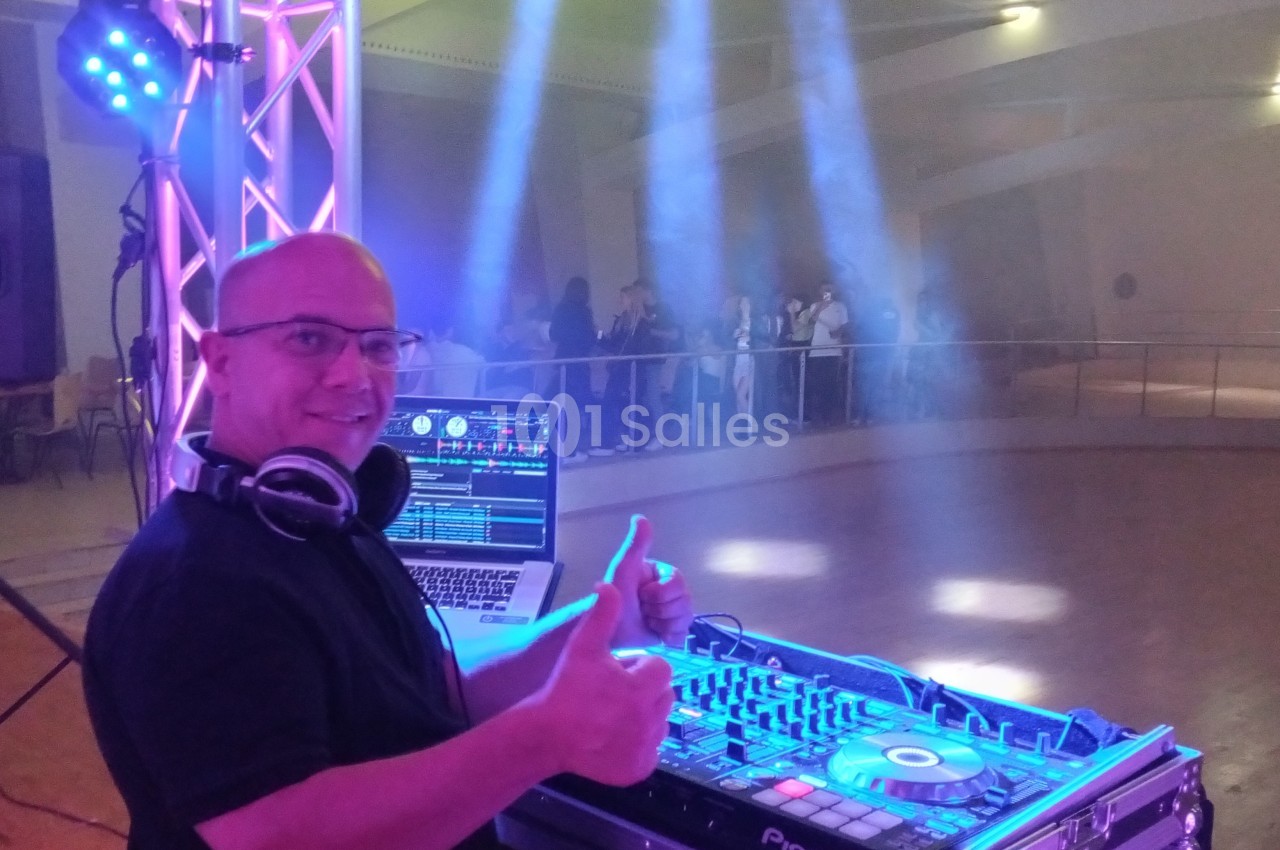 Un DJ souriant devant sa table de mixage, avec des lumières colorées et une piste de danse en arrière-plan.