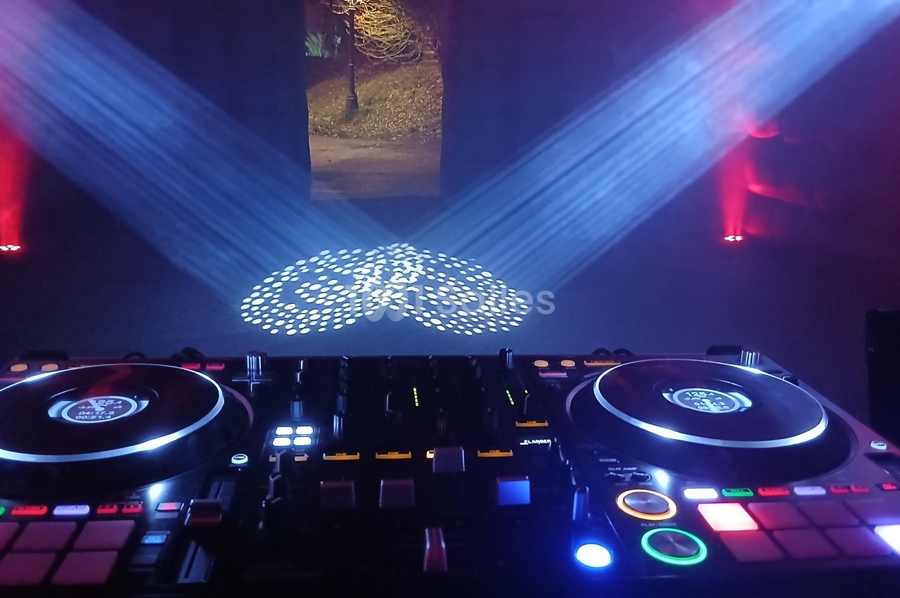 Console de mixage DJ éclairée avec des projecteurs colorés et motifs lumineux au sol dans une ambiance sombre.