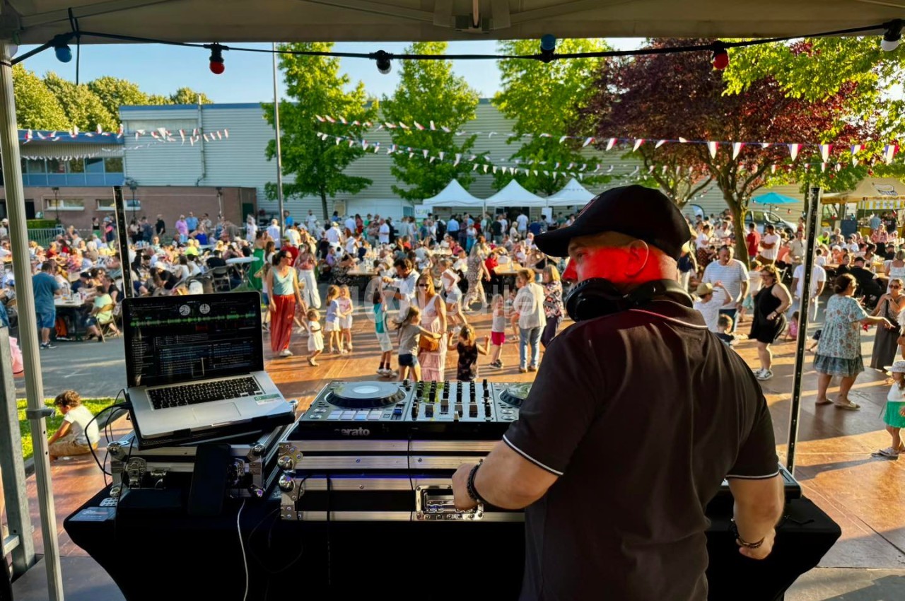 Un DJ mixe sur une scène en plein air, face à une foule rassemblée lors d'un événement festif en journée.