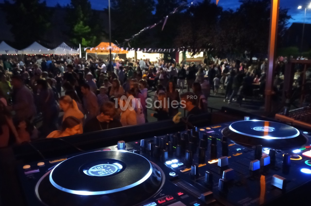 Vue d'une table de mixage en premier plan avec une foule rassemblée lors d'un événement en plein air, de nuit.