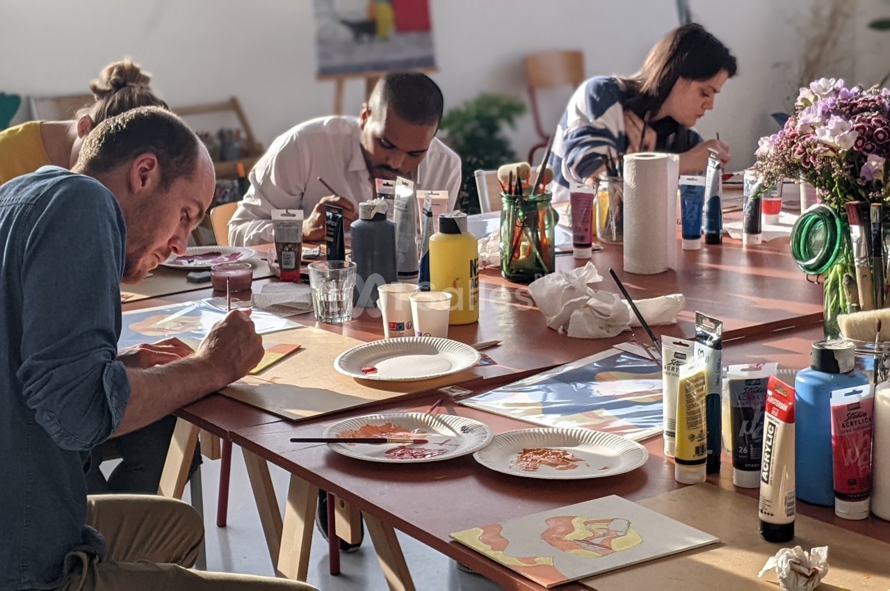 Des personnes concentrées peignent sur des feuilles autour d'une table, entourées de matériel artistique.