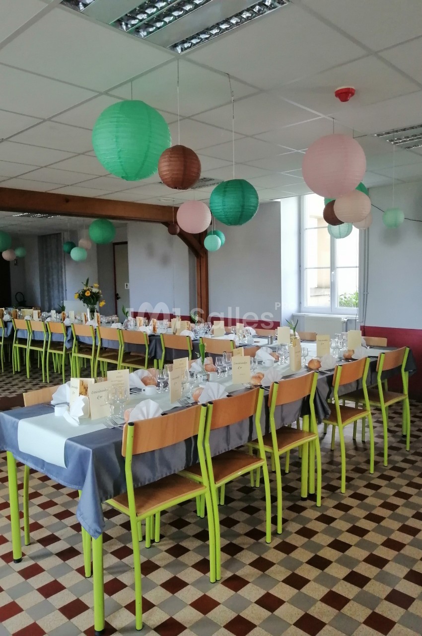 Salle décorée pour un événement, avec tables alignées, nappes grises, chaises vertes et lanternes suspendues colorées.