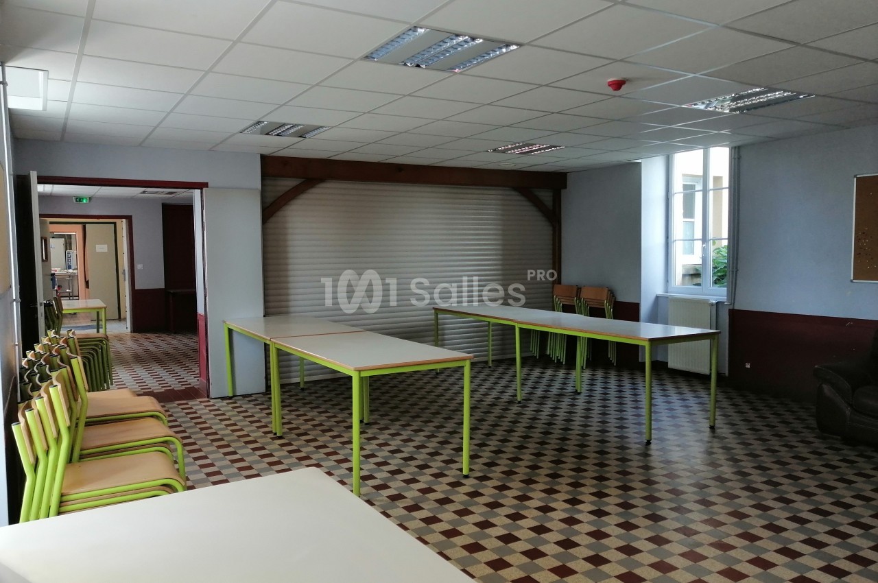 Salle vide avec tables et chaises empilées, sol en carrelage à motifs et lumière naturelle entrant par les fenêtres.