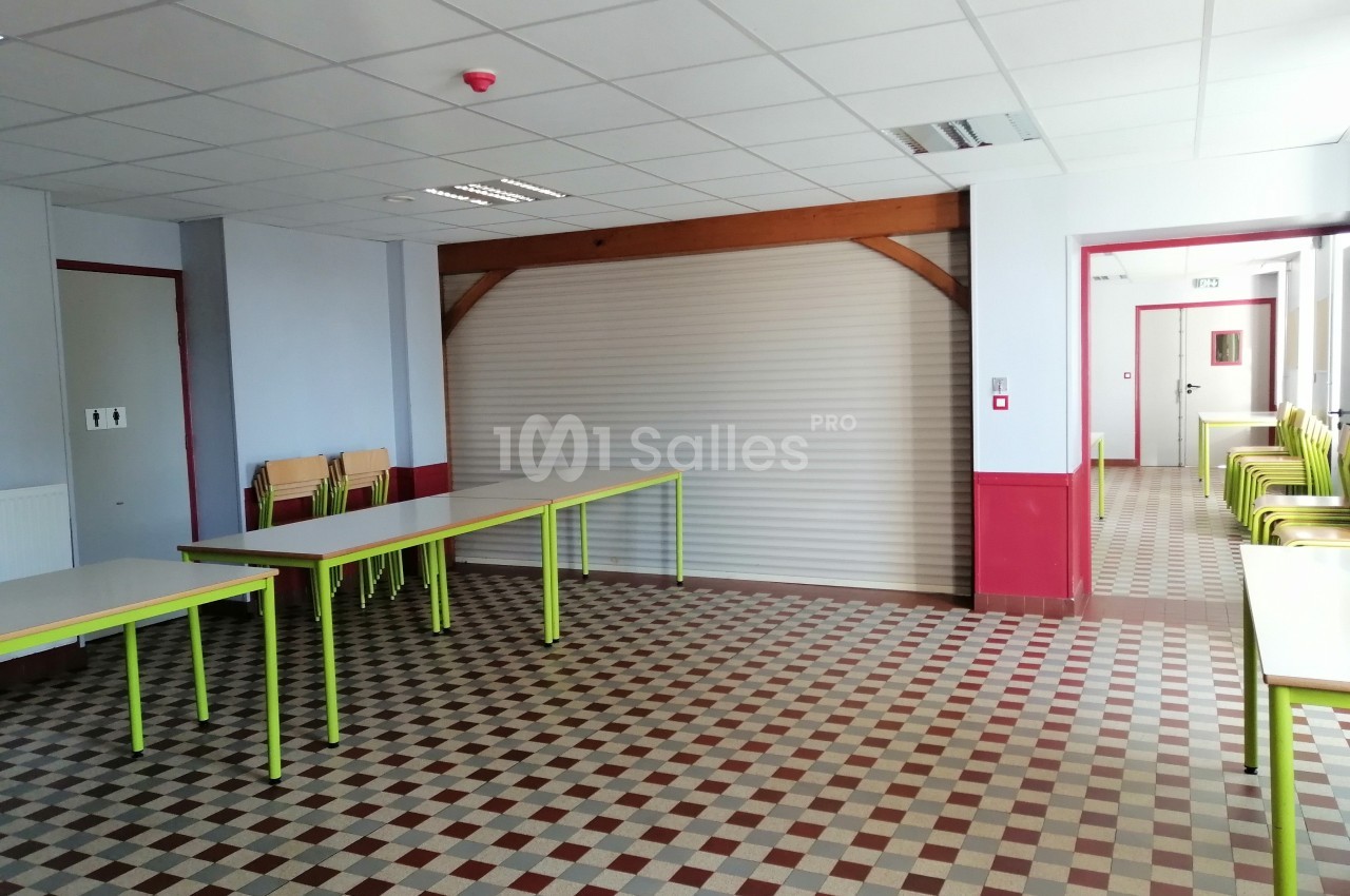 Salle vide avec tables et chaises empilées, sol à carreaux, murs clairs et rideau métallique fermé au fond.