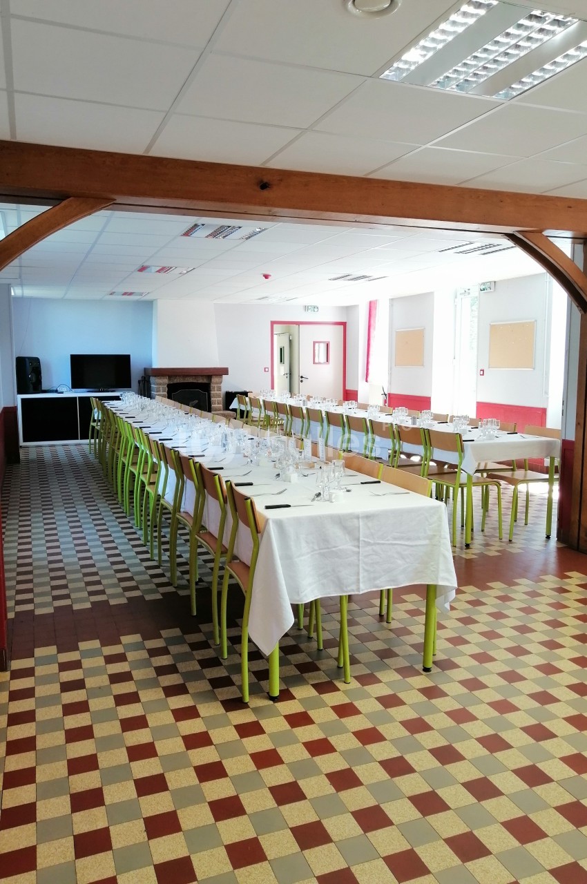 Salle lumineuse avec tables alignées, nappes blanches, chaises vertes et sol à carreaux, prête pour un événement.