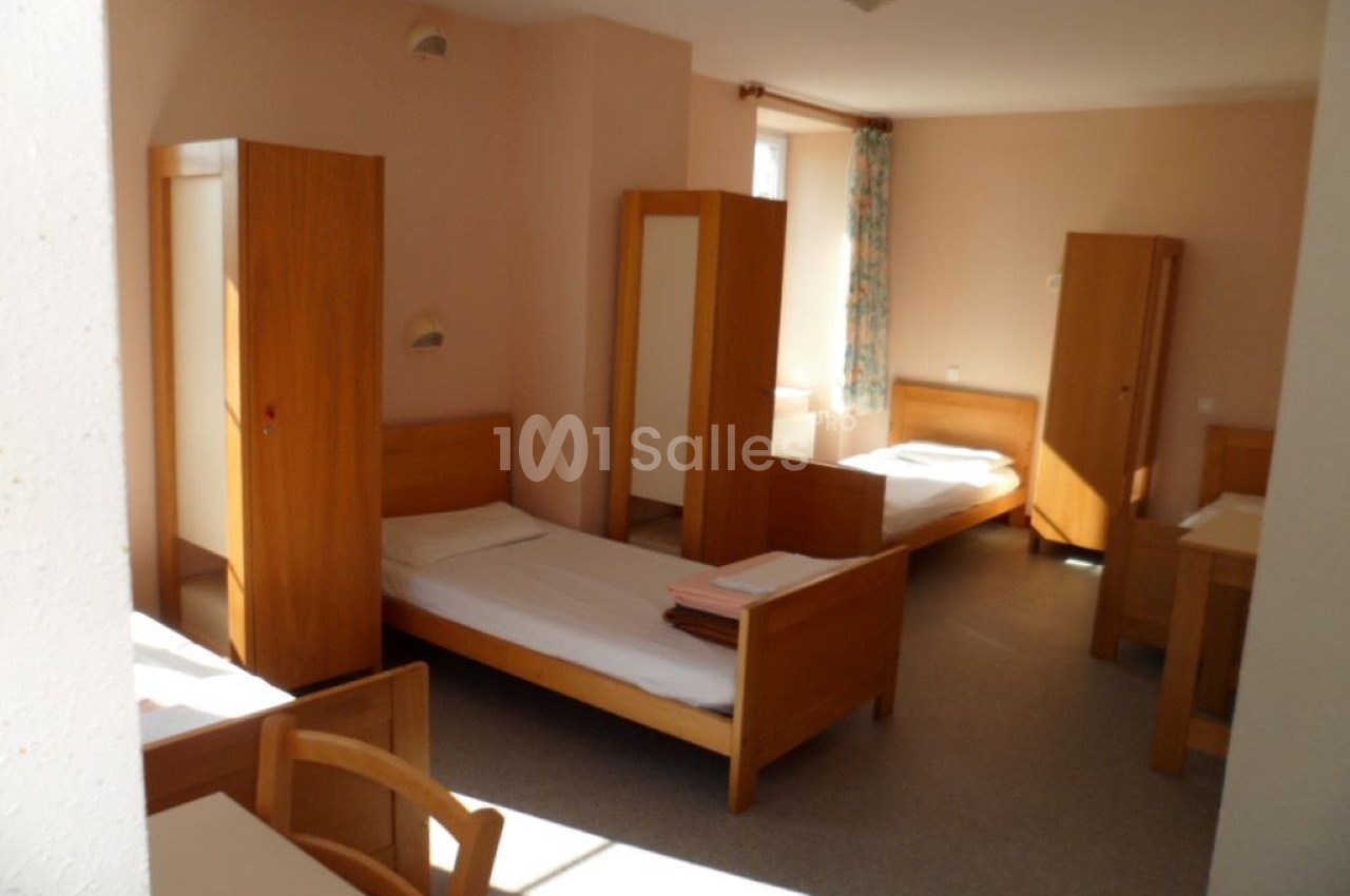 Chambre lumineuse avec plusieurs lits simples, armoires en bois et une fenêtre avec rideaux fleuris.