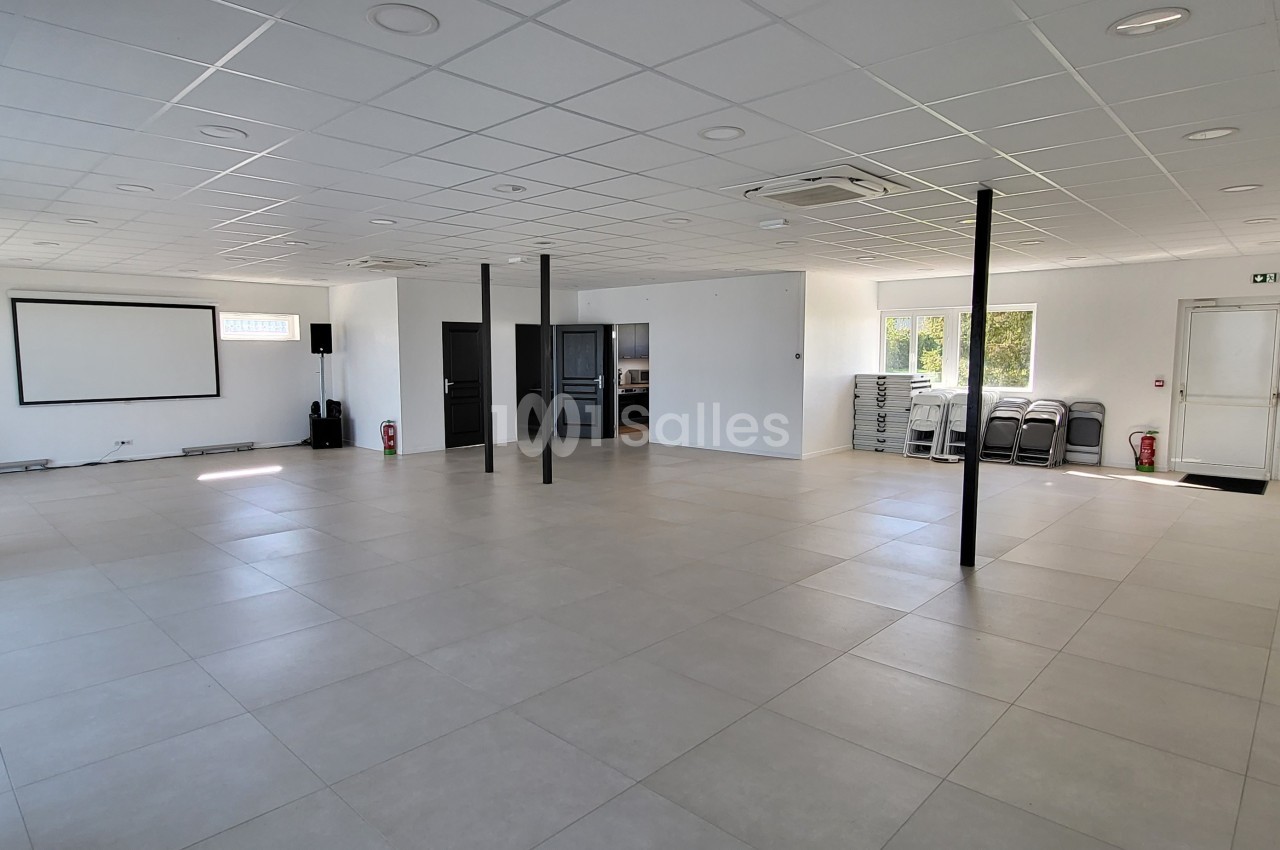 Salle vide lumineuse avec carrelage clair, murs blancs, écran de projection, chaises empilées et baies vitrées.