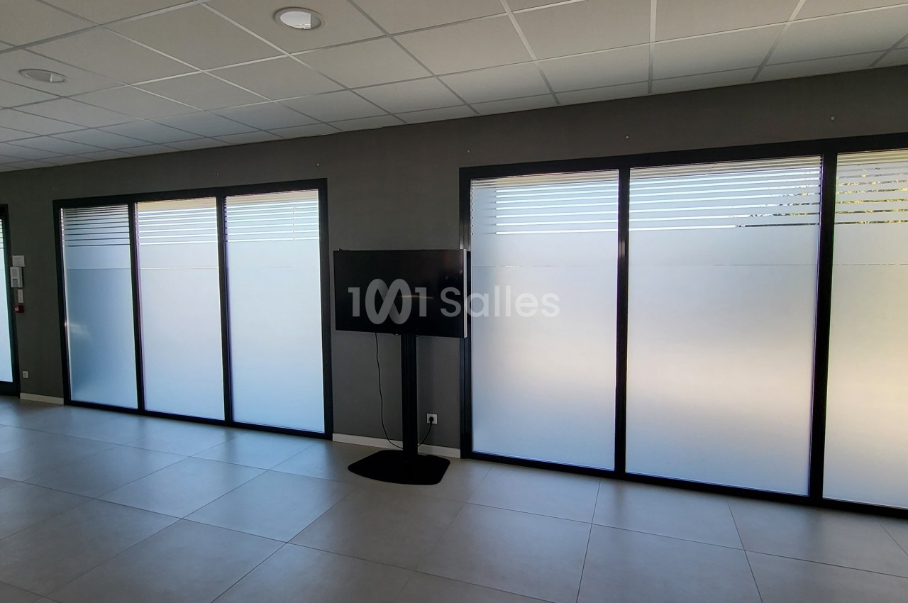 Salle lumineuse avec grandes baies vitrées partiellement opaques, sol en carrelage clair et écran sur pied.